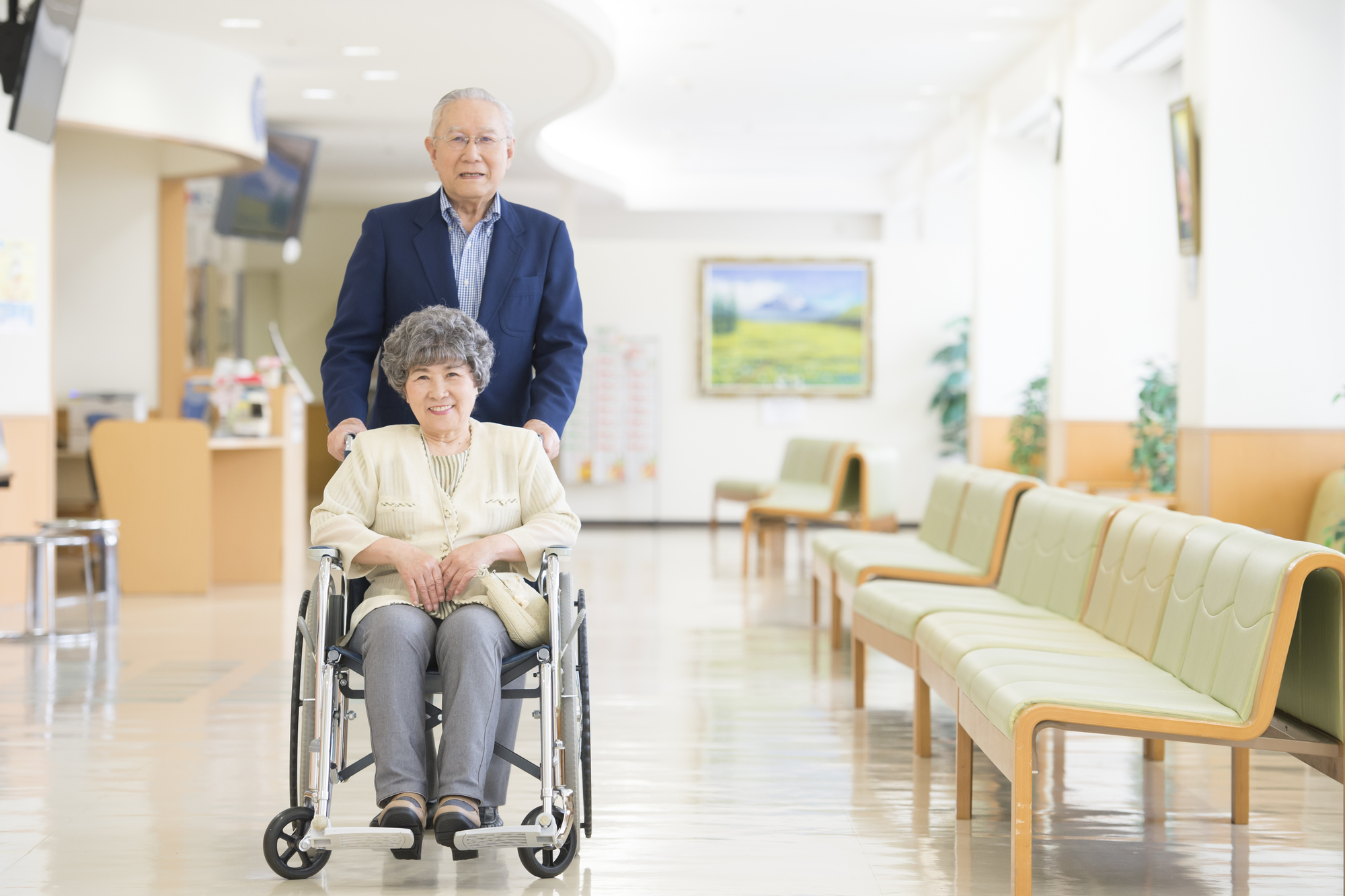 病院の待合室で車椅子に座っている高齢の女性とそれを押す高齢の男性